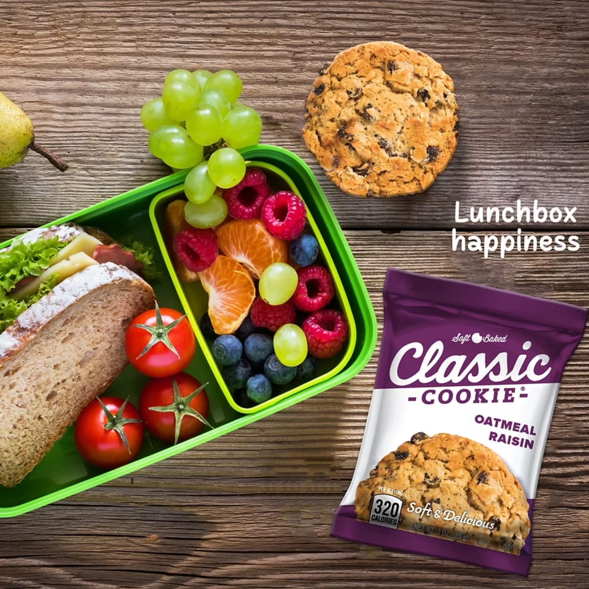 Lunchbox with sandwich, fruits, and vegetables next to a cookie and a package of classic cookie oatmeal raisin on a wooden surface.