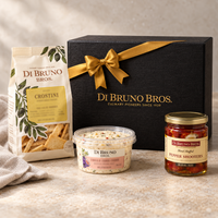 Black Di Bruno Bros. Happy Hour Kit gift box beside crostini, a cheese spread, and pepper shooters. Styling props: tabletop and linen. The gold ribbon is part of the referenced gift presentation.
