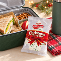 Individually wrapped Little Debbie Vanilla Christmas Tree Cake beside an open lunchbox with unbranded lunch items.